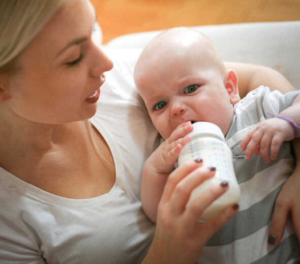 Mama trzyma bobasa na rękach, karmi go butelką i uspokaja niemowlę mówiąc do niego. Dziecko trzyma butelkę jedną rączką i patrzy na wprost gryząc smoczek od butelki.