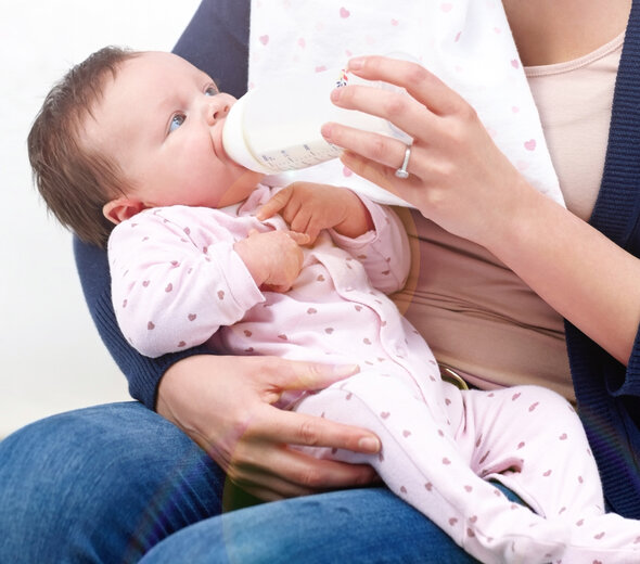 Dziecko siedzi na kolanach mamy i jest podtrzymywane pod plecami i główką ręką rodzica. Niemowlę pije mleko z butelki do karmienia i patrzy ze spokojem na mamę.