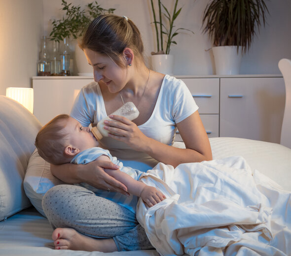 Mama trzyma dziecko na rękach siedząc ze skrzyżowanymi nogami na łóżku. Niemowlę pije mleko z butelki do karmienia, rączki ma ułożone wzdłuż tułowia i spokojnie pryzmuje pokarm.