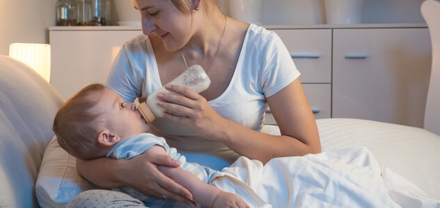 Mama trzyma dziecko na rękach siedząc ze skrzyżowanymi nogami na łóżku. Niemowlę pije mleko z butelki do karmienia, rączki ma ułożone wzdłuż tułowia i spokojnie pryzmuje pokarm.