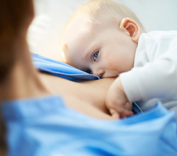 Mama siedzi i podtrzymuje przy piersi swoje dziecko. Dziecko wtulone w pierś rodzicielki ochoczo wypija naturalne mleko matki patrząc na kobietę szeroko otwartymi, dużymi, niebieskimi oczami.