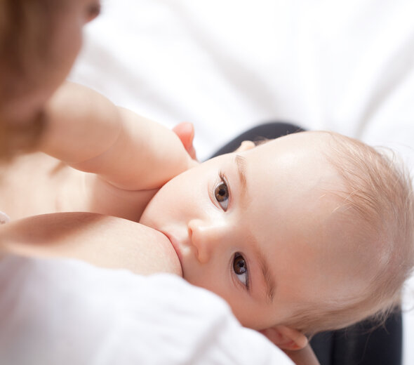 Niemowlę przytulone do klatki piersiowej mamy obejmuje ustami cały sutek rodzicielki i ssie naturalne mleko matki. Dziecko interesuje się mamą podczas jedzenia, zaczepiając jej włosy rączką.