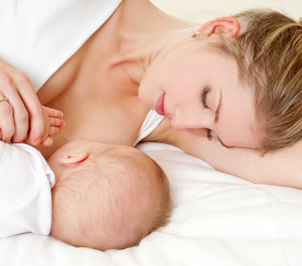 Mama leży na lewym boku zwrócona do leżącego obok niemowlęcia przystawia go do piersi w celu podania naturalnego pokarmu jakim jest mleko matki. Dziecko ze spokojem trzyma rękę kobiety.