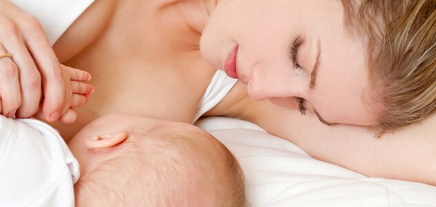 Mama leży na lewym boku zwrócona do leżącego obok niemowlęcia przystawia go do piersi w celu podania naturalnego pokarmu jakim jest mleko matki. Dziecko ze spokojem trzyma rękę kobiety.