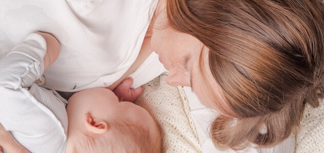 Mama leży na boku, na łóżku i przystawia nabrzmiałą pierś do ust leżącego przy niej niemowlaka w celu podania naturalnego mleka matki. Dziecko rączką kurczowo przytrzymuje mamę za bluzkę.