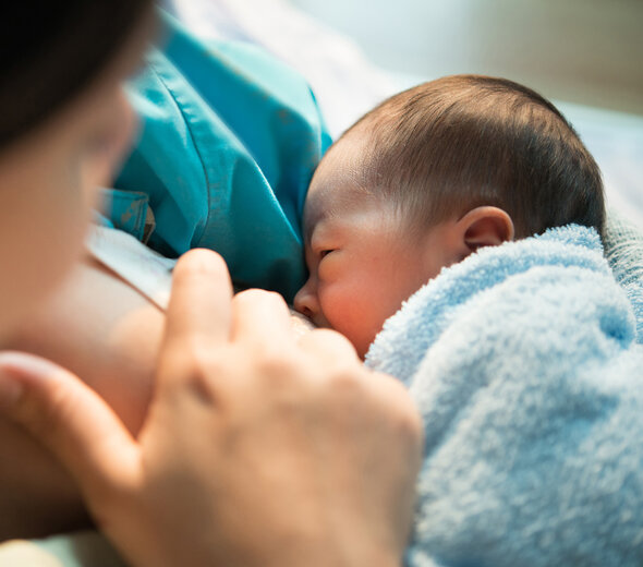 Mama siedzi i przytula do klatki piersiowej niemowlę. Przystawione dziecko do piersi pije naturalny pokarm matki. Noworodek wtula się w pierś kobiety i chętnie zjada posiłek.