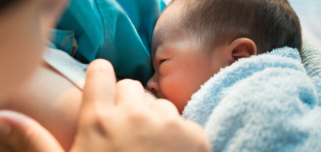 Mama siedzi i przytula do klatki piersiowej niemowlę. Przystawione dziecko do piersi pije naturalny pokarm matki. Noworodek wtula się w pierś kobiety i chętnie zjada posiłek.