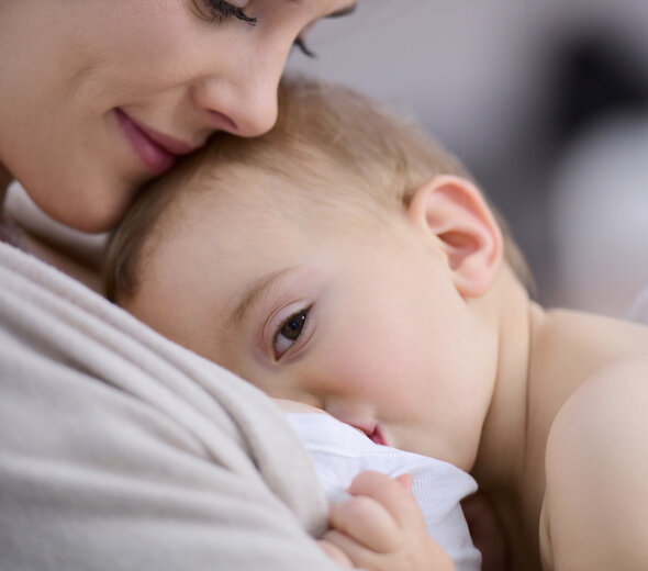 Mama przytula do piersi dziecko, które przystawiła do piersi w celu podania pożywnego mleka matki. Spokojne, niemowlę z otwartymi oczami zjada pokarm.