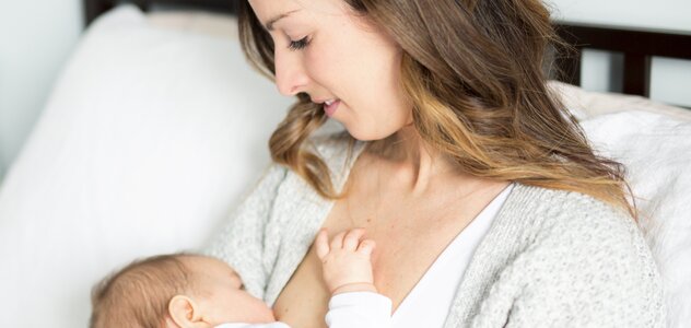 Mama siedzi na łóżku i obejmuje w ramionach noworodka, którego przystawia do piersi aby podać mu najpożywniejszy pokarm jakim jest naturalne mleko matki.