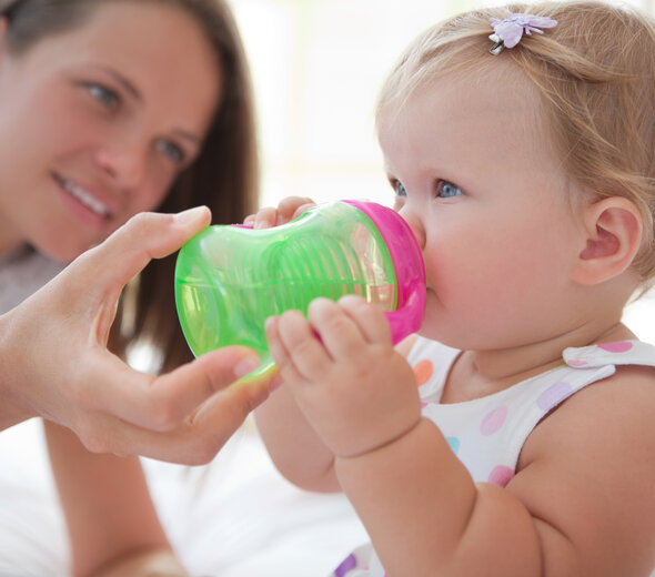 Mama podtrzymuje dziecku kubek niekapek przy buzi. Dziewczynka trzyma rączkami i pije samodzielnie napój, z niewielką asekuracją kobiety leżącej obok dziecka na łóżku.