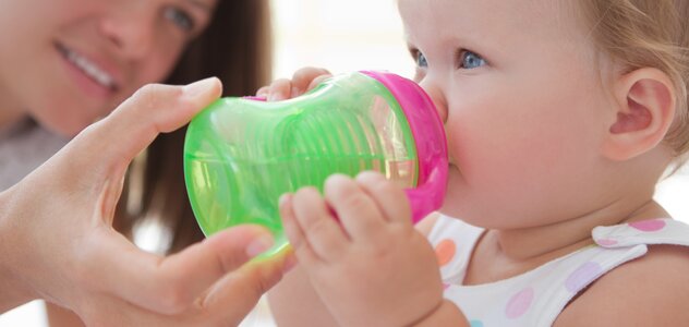 Mama podtrzymuje dziecku kubek niekapek przy buzi. Dziewczynka trzyma rączkami i pije samodzielnie napój, z niewielką asekuracją kobiety leżącej obok dziecka na łóżku.