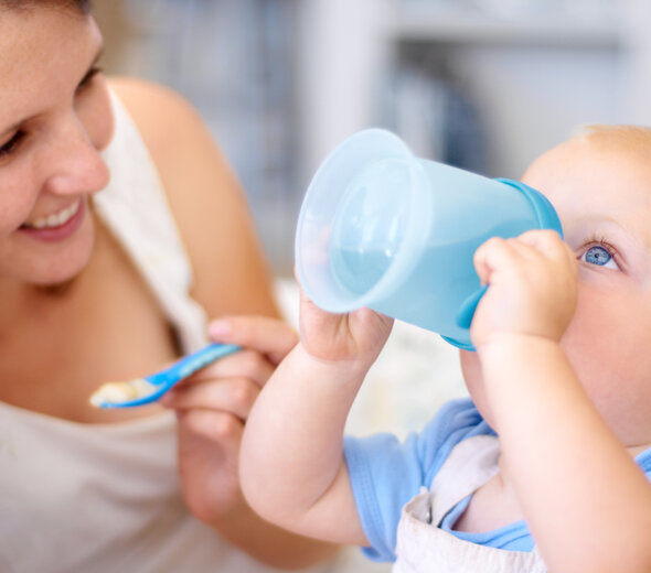 Uśmiechnięta mama podaje pokarm łyżeczką dziecku. Niemowlę samodzielnie trzyma w rączkach kubek niekapek i pije z niego wodę, która jest podstawowym elementem diety.