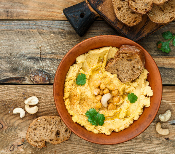 W misce znajduje się hummus posypany ciecierzycą, pietruszką i nerkowcami. Obok, na desce leżą kromki chleba. Danie znajduje się na drewnianym stole.