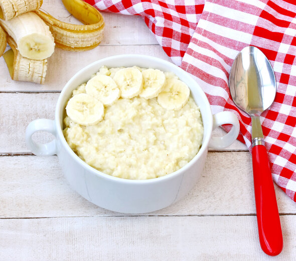 W białej miseczce znajduje się owsianka oraz plasterki banana. obok leży łyżka z czerwoną rączką oraz ściereczka w kratę. Posiłek znajduje się na stole z białego drewna.