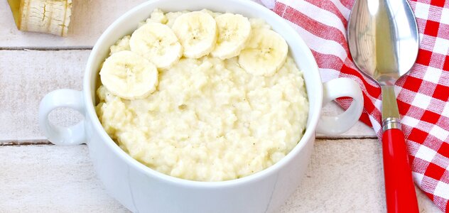 W białej miseczce znajduje się owsianka oraz plasterki banana. obok leży łyżka z czerwoną rączką oraz ściereczka w kratę. Posiłek znajduje się na stole z białego drewna.