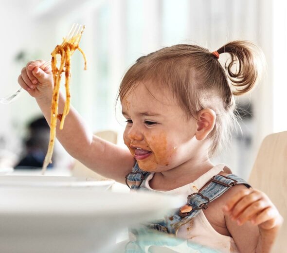 Mała dziewczynka siedzi w foteliku do karmienia i je spaghetti. Ma dżinsy ogrodniczki i białą bluzeczkę. Ma włosy spięte w kitkę. Jest cała umorusana. Bawi się jedzeniem.