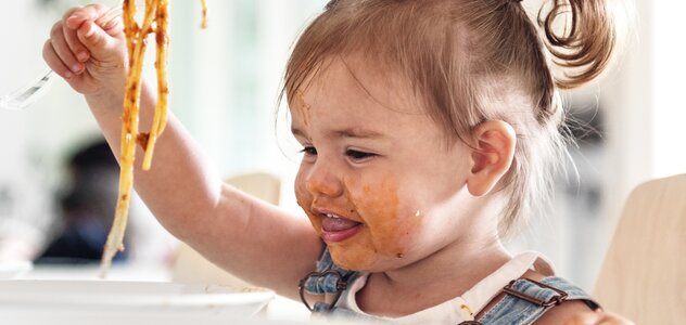 Mała dziewczynka siedzi w foteliku do karmienia i je spaghetti. Ma dżinsy ogrodniczki i białą bluzeczkę. Ma włosy spięte w kitkę. Jest cała umorusana. Bawi się jedzeniem.