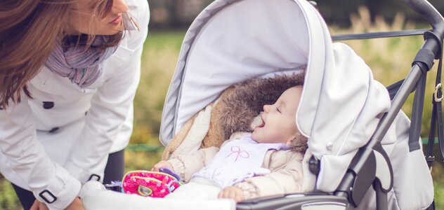 Mama wyszła na spacer ze swoim dzieckiem. Ubrana w ciepłą kurtkę kobieta zagląda do wózka w którym siedzi niemowlę. Bobas uśmiecha się szeroko na widok rodzica.