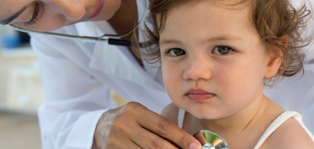 Dziewczynka z niezbyt szczęśliwa miną siedzi na fotelu w gabinecie lekarskim i klatka piersiowa jest osłuchiwana stetoskopem przez Panią doktor pediatrę.