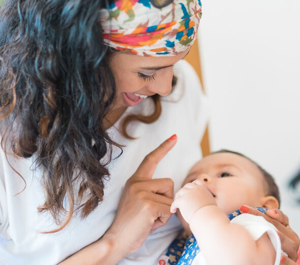 Uśmiechnięta mama w kolorowej chustce zabawia swoją córeczkę, która leży na jej ramieniu i uważanie wpatruje się w każdy ruch kobiety. Dziecko jest bardzo zainteresowane palcem mamy.