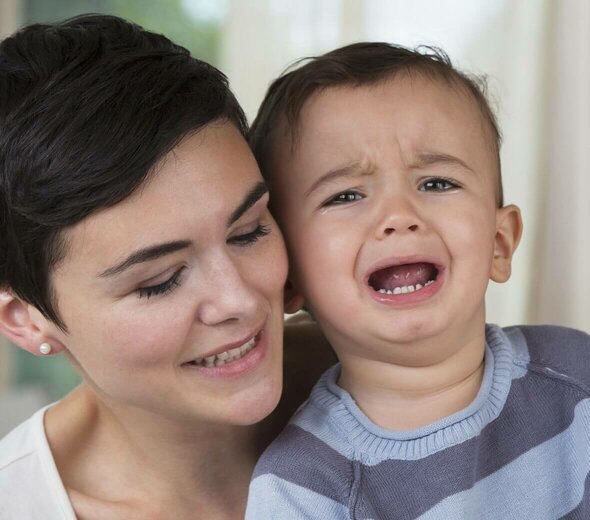 Mama stoi za synkiem i przytula go od tyłu. Dziecko ma łzy w oczach i otwartą z bólu buzię. Bóle wzrostowe u dzieci bywają bardzo przykrym doświadczeniem zarówno dla dziecka jak i rodzica.