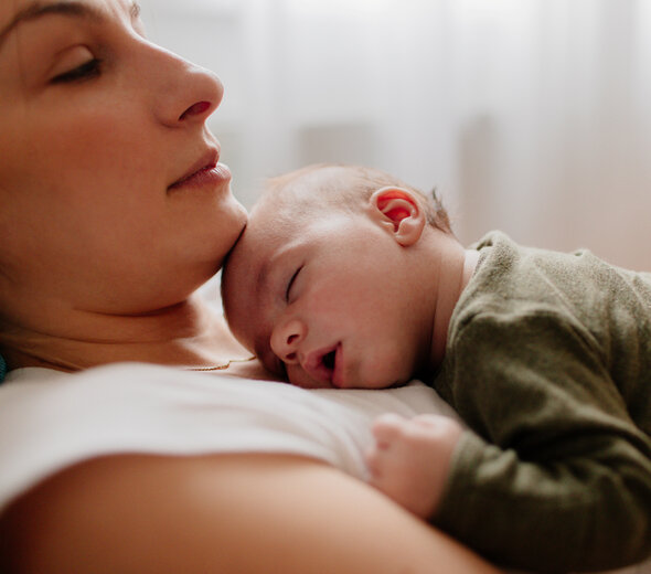 Spokojna mama z zamkniętymi oczami leży na łóżku podpierając głowę o zagłówek. Niemowlę śpi na jej klatce piersiowej z szeroko otwartą buzią. Bliskość jest bardzo istotna dla dziecka.