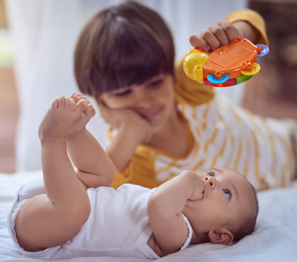 Rodzeństwo bawi się leżąc na łóżku. Niemowlę leży na pleckach i z zaciekawieniem patrzy na grzechotkę, którą nad jego twarzą porusza starszy brak leżący obok.