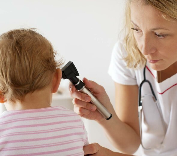 Kilkunastomiesięczne dziecko siedzi samodzielnie w gabinecie lekarskim i spokojnie poddaje się badaniu lekarskiemu. Pani doktor zagląda mu do ucha sprawdzając stan zdrowia.