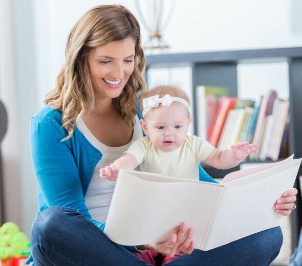 Mama z szerokim uśmiechem siedzi ze skrzyżowanymi nogami na podłodze i trzyma przed sobą i dzieckiem książkę. Niemowlę siedzi na kolanach i jest bardzo zainteresowane lekturą. 