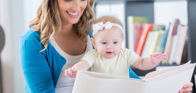 Mama z szerokim uśmiechem siedzi ze skrzyżowanymi nogami na podłodze i trzyma przed sobą i dzieckiem książkę. Niemowlę siedzi na kolanach i jest bardzo zainteresowane lekturą. 