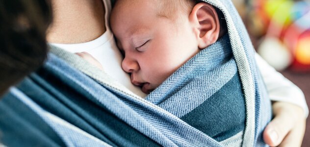 Mama chustuje niemowlę. Dziecko starannie owinięte długą, przeznaczona do tej czynności chustą smacznie sobie śpi wyczuwając  bliskość mamy i słysząc jej bicie serca.