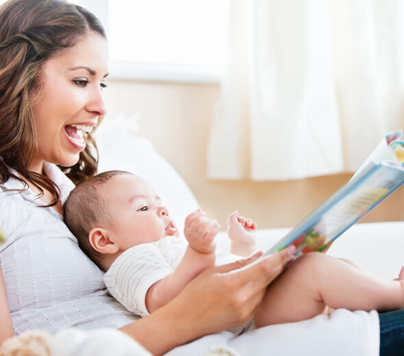 Mama z szerokim uśmiechem na twarzy trzyma leżące  na kolanach do niej tyłem niemowlę. Kobieta pokazuje noworodkowi kolorową książkę. Dziecko jest bardzo zainteresowane.