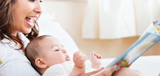 Mama z szerokim uśmiechem na twarzy trzyma leżące  na kolanach do niej tyłem niemowlę. Kobieta pokazuje noworodkowi kolorową książkę. Dziecko jest bardzo zainteresowane.