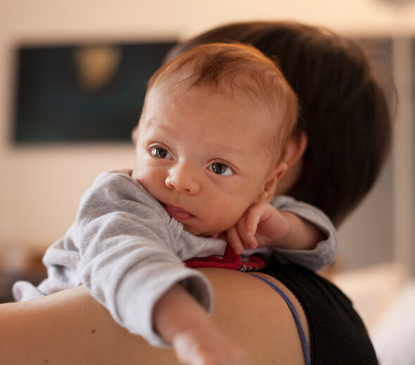 Mama trzyma wysoko ponad ramieniem niemowlę, opierając je brzuszkiem o swoje ramię. Noworodek unosi wysoko gółkę z zaciekawieniem rozglądając się po pokoju.