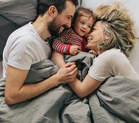 Szeroko uśmiechnięta rodzinka wyleguje się w łóżku. Tata z lewej strony zwrócony w stronę rodziny, mama z prawej strony wtula się w synka leżącego pomiędzy rodzicami.