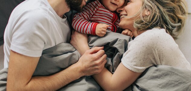 Szeroko uśmiechnięta rodzinka wyleguje się w łóżku. Tata z lewej strony zwrócony w stronę rodziny, mama z prawej strony wtula się w synka leżącego pomiędzy rodzicami.