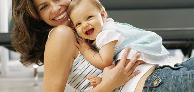 Mama w pozycji półleżącej wspiera się łokciami na dywanie, na brzuchu siedzi roześmiana córeczka. Zabawy z niemowlakiem na dywanie to jedno z najprzyjemniejszych zajęć.