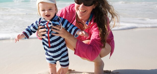 Młoda mama razem ze swoim kilkumiesięcznym dzieckiem przebywa na plaży. Mały bobas ubrany w pasiaste body oraz czapeczkę świetnie bawi się chodząc z pomocą mamy boso po piasku. 