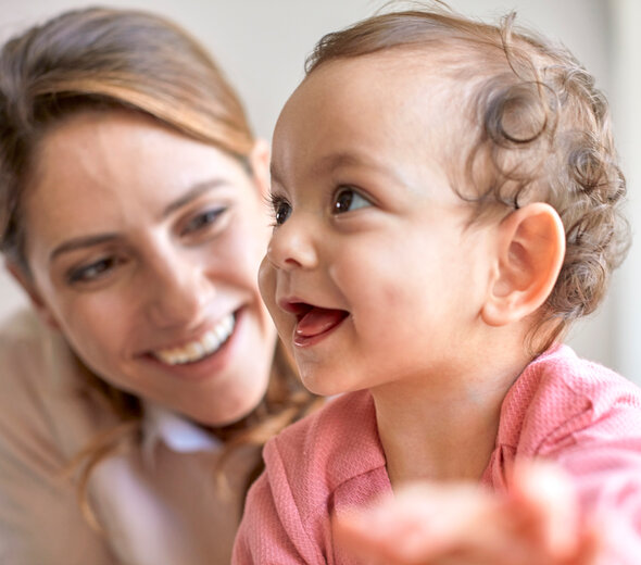 Młoda mama przebywa razem z kilkumiesięcznym dzieckiem. Małe dziecko siedzi samodzielnie na podłodze i szeroko się uśmiecha patrząc w górę. Za dziewczynką widać uśmiechniętą mamę.