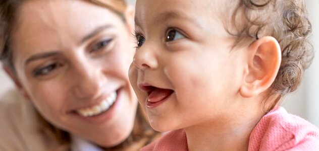 Młoda mama przebywa razem z kilkumiesięcznym dzieckiem. Małe dziecko siedzi samodzielnie na podłodze i szeroko się uśmiecha patrząc w górę. Za dziewczynką widać uśmiechniętą mamę.