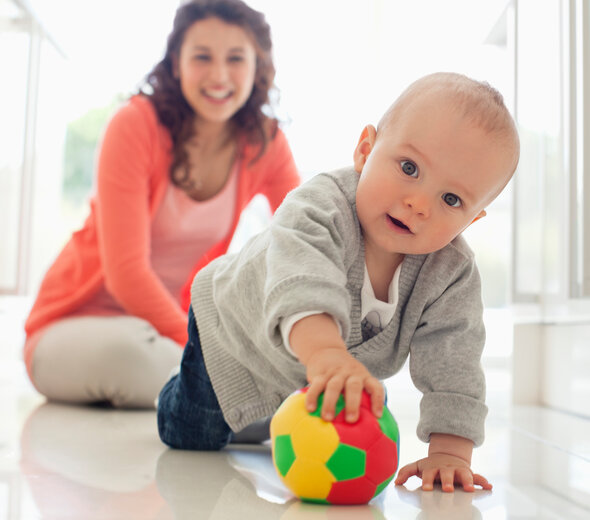 Młoda mama razem z kilku miesięcznym bobasem przebywają w kuchni. Rodzicielka z szerokim uśmiechem siedzi na podłodze i przygląda się synkowi. Niemowlę na czworakach goni piłkę.