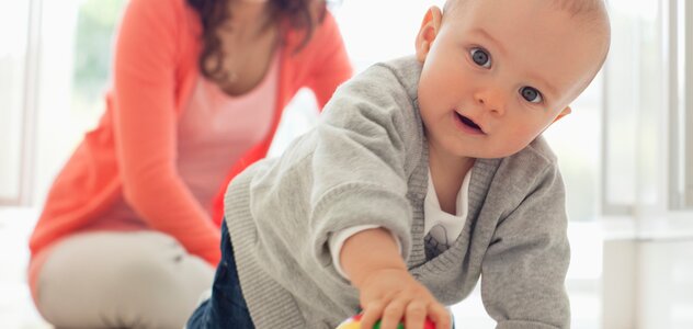 Młoda mama razem z kilku miesięcznym bobasem przebywają w kuchni. Rodzicielka z szerokim uśmiechem siedzi na podłodze i przygląda się synkowi. Niemowlę na czworakach goni piłkę.