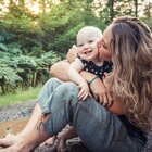 Młoda mama razem ze swoim kilku miesięcznym bobasem przebywa na świeżym powietrzu. Kobieta siedzi na ziemi w błocie, na rękach trzyma małego noworodka i całuje go w policzek.