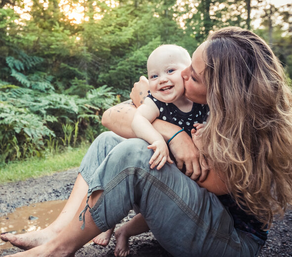 Młoda mama razem ze swoim kilku miesięcznym bobasem przebywa na świeżym powietrzu. Kobieta siedzi na ziemi w błocie, na rękach trzyma małego noworodka i całuje go w policzek.