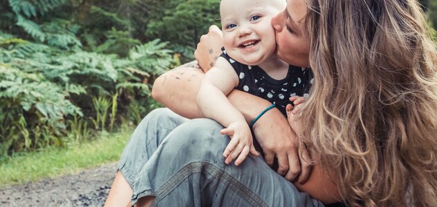 Młoda mama razem ze swoim kilku miesięcznym bobasem przebywa na świeżym powietrzu. Kobieta siedzi na ziemi w błocie, na rękach trzyma małego noworodka i całuje go w policzek.