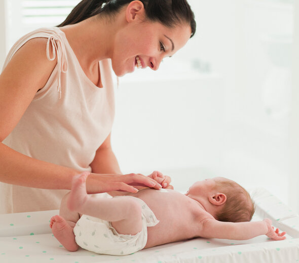 Szczęśliwa młoda mama z szerokim uśmiechem przebywa w pokoiku dziecka razem ze swoim niedawno narodzonym niemowlakiem. Bobas leży na przewijaku, a mama smaruje go kremem.