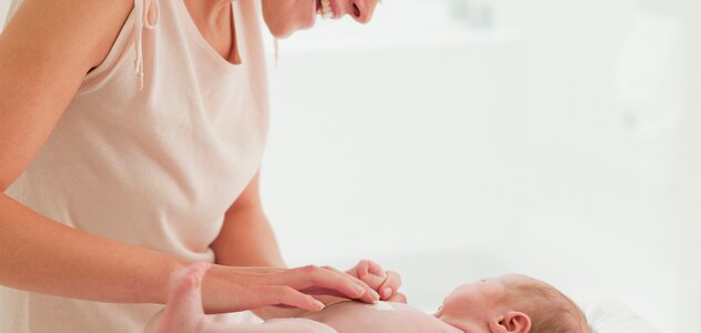 Szczęśliwa młoda mama z szerokim uśmiechem przebywa w pokoiku dziecka razem ze swoim niedawno narodzonym niemowlakiem. Bobas leży na przewijaku, a mama smaruje go kremem.