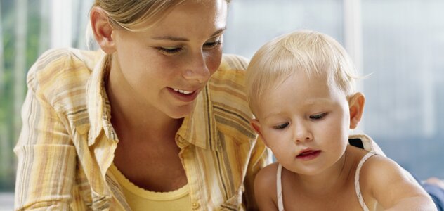 Młoda mama przebywa razem ze swoim kilku miesięcznym bobasem w salonie. Rodzicielka leży na włochatym dywanie. Obok kobiety na brzuchu leży dziecko i ogląda książeczkę.