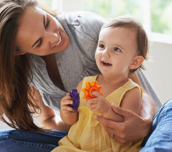 Młoda mama siedzi na podłodze w salonie. Kobieta nachyla się i spogląda na kilku miesięczną córkę, która siedzi tuż przed nią. Dziewczynka ubrana jest w żółtą sukienkę i trzyma kolorowe klocki w rękach.