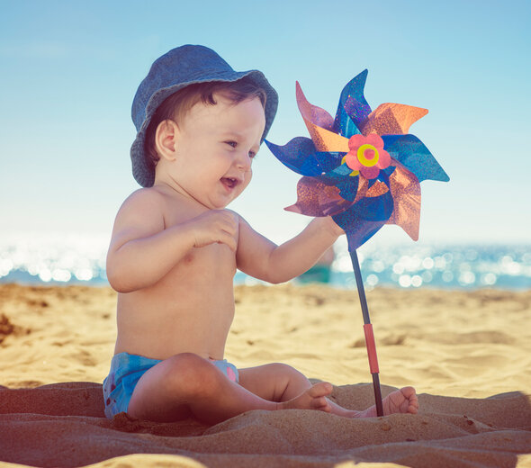 Kilku miesięczny bobas samodzielnie siedzi na plaży. Dziecko ubrane w pieluszkę i kapelusik siedzi na piasku w cieniu parasola. Niemowlę bawi się wiatrakiem, który wbity jest w ziemię.